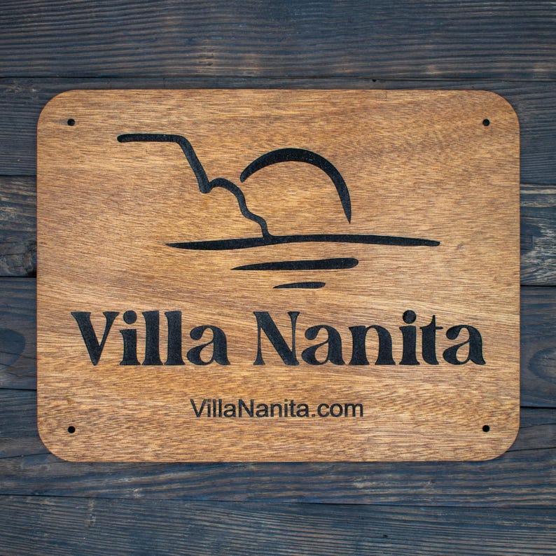 May include: A wooden sign with the text "Villa Nanita" and the website address "VillaNanita.com". The sign features a stylised image of a sunset over water.