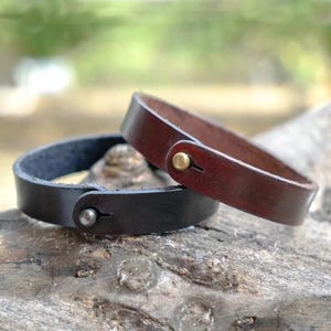 Può includere: Vengono mostrati due braccialetti in pelle, uno marrone scuro e uno nero. I braccialetti hanno un design semplice con chiusura a scatto. Il braccialetto marrone scuro ha un bottone a pressione color oro, mentre quello nero ha un bottone a pressione color argento.