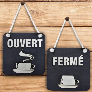 Peut inclure: Deux pancartes noires en ardoise avec des poignées en corde blanche. La pancarte de gauche indique "OUVERT" et présente une silhouette blanche d'une tasse de café avec de la vapeur. La pancarte de droite indique "FERMÉ" et présente une silhouette blanche d'une tasse de café.