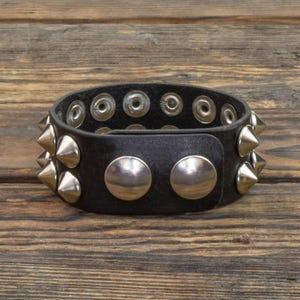 Puede incluir: Brazalete de cuero negro con tachuelas cónicas plateadas y grandes remaches plateados. El brazalete tiene un cierre de botón a presión. Accesorio de estilo punk rock o motero, sobre fondo de madera.