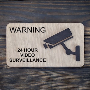 以下が含まれることがあります： 「WARNING 24 HOUR VIDEO SURVEILLANCE」という文字と、黒いシルエットの防犯カメラが描かれた木製の看板です。