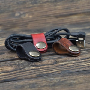 May include: Three leather cable organizers in black, brown, and red with brass accents. The organizers are designed to hold and manage cords and cables.
