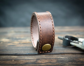 May include: A brown leather bracelet with a brass button closure. The bracelet is stitched with brown thread.
