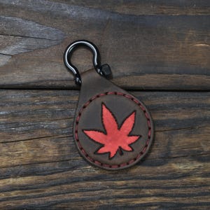 Puede incluir: Un llavero de cuero marrón con un diseño de hoja de arce roja cosido en un parche circular. El llavero tiene un cierre de metal negro y costuras rojas. Fondo de superficie de madera oscura.