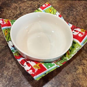 May include: A white ceramic bowl rests on a colorful, square bowl cozy. The cozy features a festive design with red, green, and white elements, including cartoon characters. The bowl is placed on a brown countertop.