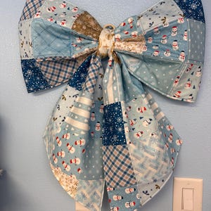 May include: A large decorative bow made from quilted fabric featuring a patchwork design of light blue, dark blue, and white patterns. The fabric displays various winter-themed prints including snowmen, snowflakes, and plaid, with a central knot.