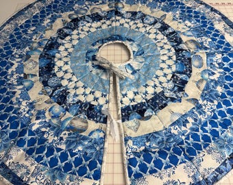 May include: A round, quilted Christmas tree skirt in shades of blue and white. The skirt features a variety of patterns, including floral designs, geometric shapes, and Christmas ornaments. It has a split opening with ties for easy placement around a tree.