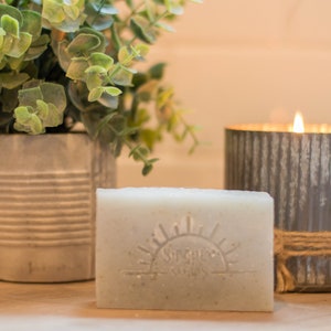 May include: A white bar of soap with a sun and the words "Sunrise Soaps" embossed on it. The soap is sitting on a wooden surface next to a gray metal candle holder with a lit candle inside. A potted plant with green leaves is in the background.