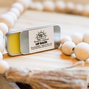 Może przedstawiać: Otwarta metalowa puszka z balsamem do ust z żółtym balsamem w środku, z napisem "Simple Goods Lip Balm". Etykieta zawiera składniki i słowa "Peppermint Dandelion Infused Lip Balm". W tle girlanda z drewnianych koralików i frędzel.