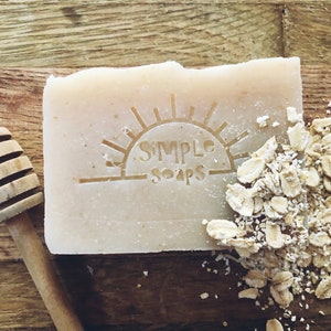 May include: A bar of soap with a sun and the words "Simple Soaps" printed on it. The soap is on a wooden surface with a honey dipper and a pile of oatmeal.