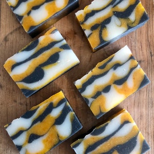 May include: Six bars of handmade soap with a black, white, and yellow swirl pattern. The soap bars are arranged on a wooden surface.