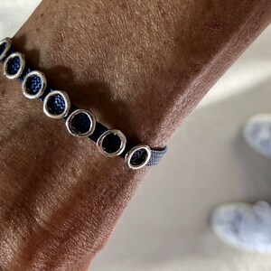 May include: A dark blue bracelet with silver-colored circular accents. The bracelet is worn on a person's wrist. The bracelet's design features a woven band with evenly spaced, circular silver-toned embellishments.