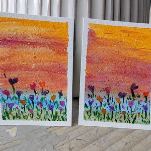 May include: Two watercolor paintings of a sunset over a field of flowers. The sky transitions from orange to red, with a speckled texture. The flowers are purple, pink, and orange, with green stems and leaves, set against a blue and green base.