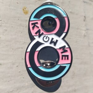 Könnte beinhalten: Eine schwarze und weiße Emaille-Anstecknadel in Form der Zahl Acht. Die Anstecknadel hat ein pinkfarbenes, blaues und weißes Streifenmuster mit den Worten "Know Me", die vertikal auf der rechten Seite der Anstecknadel stehen.