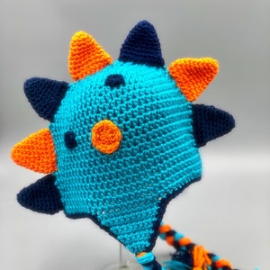 May include: A blue, orange, and navy blue crocheted hat with a long braided cord. The hat has a spiky design and a large orange circle on the front.