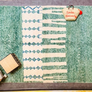 May include: A rectangular, green and white rug with a geometric design. The rug features a central white section with blue diamond and stripe patterns. A woven tote bag with the text "Berber Moroccan Rugs" hangs on the rug.
