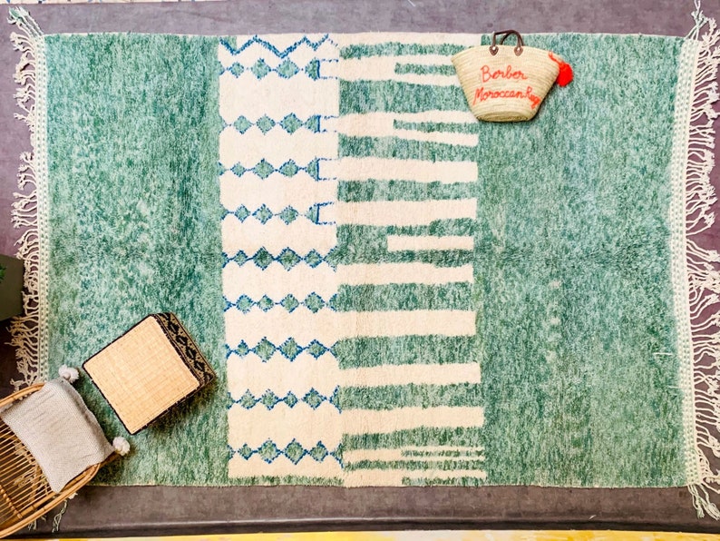 May include: A rectangular, green and white rug with a geometric design. The rug features a central white section with blue diamond and stripe patterns. A woven tote bag with the text "Berber Moroccan Rugs" hangs on the rug.