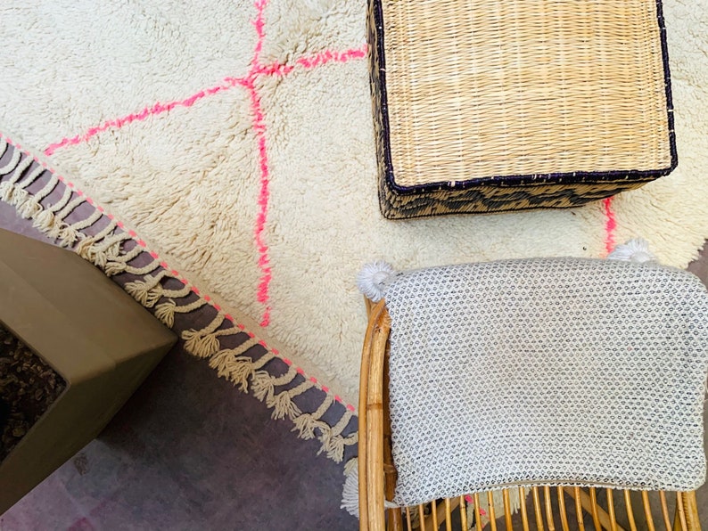 May include: A white fluffy rug with pink stitching and a woven basket with a black trim. A white woven chair with a gray cushion is in the background.