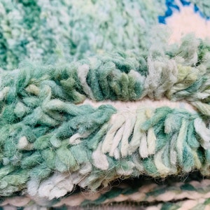 May include: Close-up of a textured rug with a shaggy pile. The rug features a blend of green, white, and blue hues, creating a soft, inviting appearance. The fibers are thick and create a plush texture, suggesting comfort and warmth.