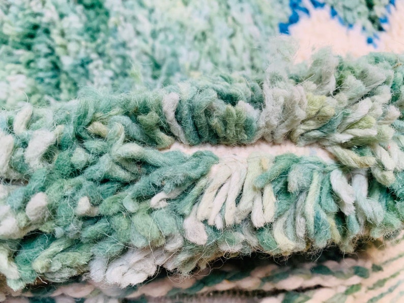 May include: Close-up of a textured rug with a shaggy pile. The rug features a blend of green, white, and blue hues, creating a soft, inviting appearance. The fibers are thick and create a plush texture, suggesting comfort and warmth.