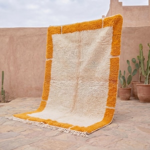 Puede incluir: Una alfombra rectangular de color blanquecino con una pila gruesa y texturizada y un borde naranja vibrante. La alfombra se exhibe al aire libre, apoyada contra una pared, con cactus y macetas de terracota al fondo. Dimensiones aproximadas: 152 cm x 229 cm.