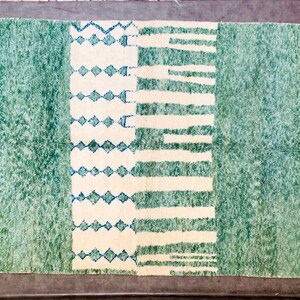 May include: A rectangular rug with a green and off-white design. The rug features a central off-white section with blue diamond and horizontal stripe patterns, flanked by green textured fields. The edges have a fringe.