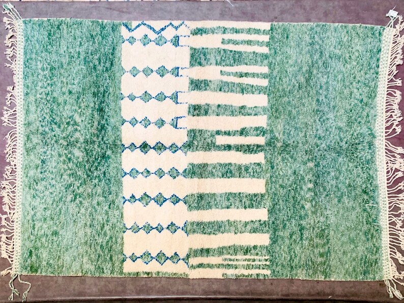 May include: A rectangular rug with a green and off-white design. The rug features a central off-white section with blue diamond and horizontal stripe patterns, flanked by green textured fields. The edges have a fringe.
