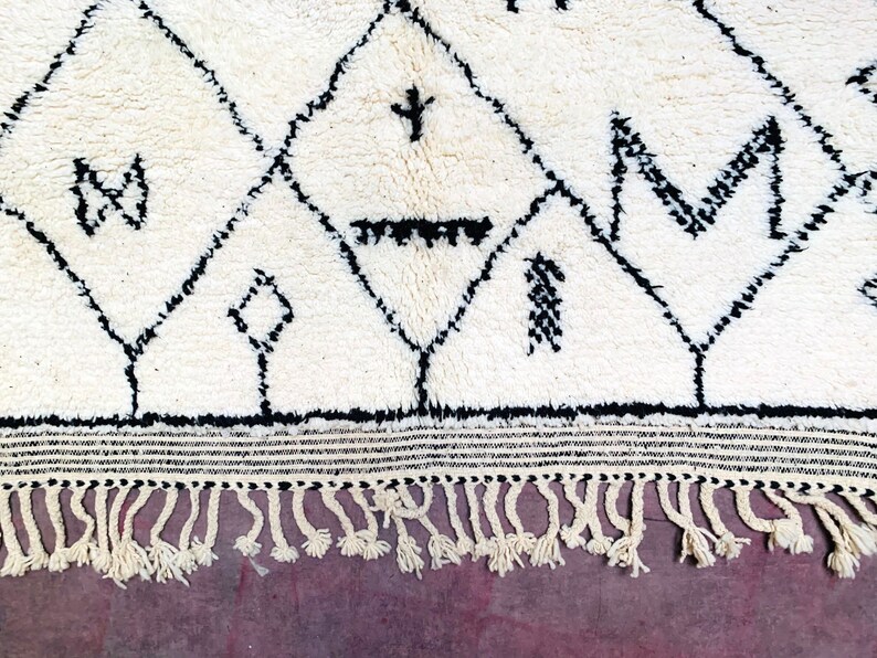 May include: A white rug with a black geometric pattern. The rug has a fringe border.