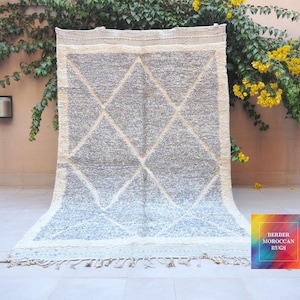May include: A rectangular Berber rug with a geometric diamond pattern in shades of grey and cream. The rug has a fringe along the bottom edge and is displayed against a neutral background with greenery and yellow flowers.