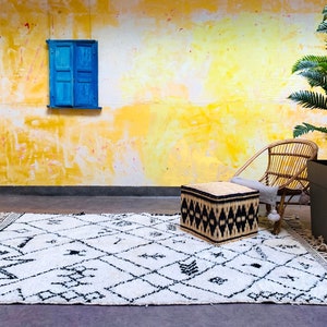 May include: A white and black patterned rug with fringe, a woven ottoman, a wicker chair, and a potted palm tree in front of a yellow wall with a blue window.