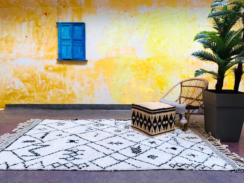 May include: A white and black patterned rug with fringe, a woven ottoman, a wicker chair, and a potted palm tree in front of a yellow wall with a blue window.