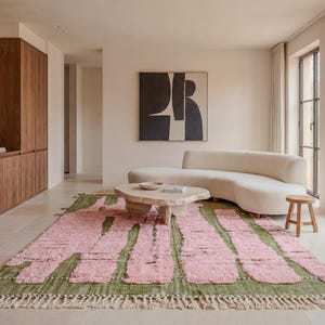 Pode incluir: Uma sala de estar moderna com um sofá curvo creme, uma mesa de centro de madeira clara e um tapete com padrões rosa e verde. A sala tem armários de madeira e uma grande pintura abstrata. Um pequeno banco de madeira fica ao lado do sofá.