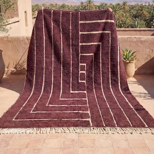 May include: A burgundy rug with a geometric design. The rug features vertical stripes and a central square pattern in cream. The rug has a fringe along the bottom edge. The rug is displayed outdoors.