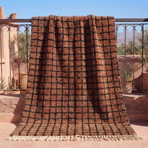 May include: A brown and black checkered rug with a fringed edge. The rug is displayed outdoors, draped over a metal railing. The rug's design features a grid pattern, and it appears to be made of a textured material.