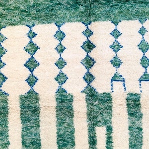 May include: A close-up of a rug with a cream-colored background and a pattern of vertical green stripes and rows of blue diamond shapes. The top of the rug has a solid green border.