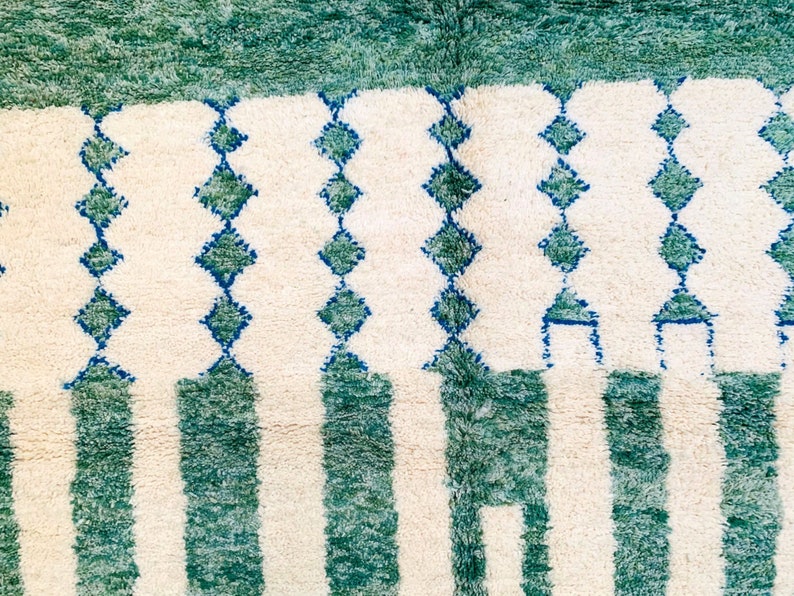 May include: A close-up of a rug with a cream-colored background and a pattern of vertical green stripes and rows of blue diamond shapes. The top of the rug has a solid green border.