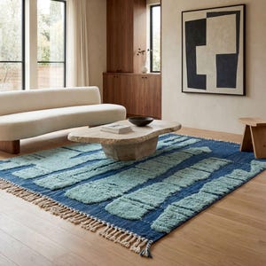 Może przedstawiać: Nowoczesny salon z niebieskim dywanem z jasnoniebieskimi geometrycznymi wzorami. Obecna jest również beżowa sofa, kamienny stolik kawowy i drewniany stołek. Pokój posiada duże okna i drewniane szafki.