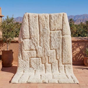 May include: A cream-colored, geometric patterned rug is displayed outdoors. The rug has a thick, textured pile and a rectangular design. The background features terracotta walls, potted plants, and a mountain range under a clear blue sky.