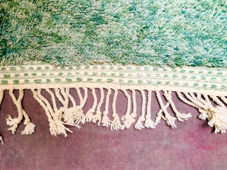 May include: Close-up of a textured rug with a teal and green color scheme. The rug features a shaggy pile and a cream-colored border with green stitching. Cream-colored fringe hangs from the bottom edge, adding a decorative element.