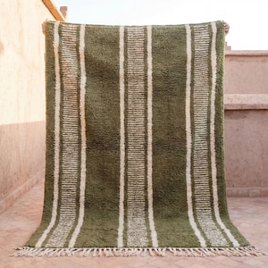 May include: A rectangular olive green rug with vertical white stripes. The rug has a shaggy texture and a fringe along the bottom edge. The rug is held up against a light-colored wall.