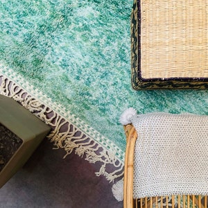May include: Overhead view of a living room setup featuring a teal shag rug with a fringed border. A woven basket with a dark patterned trim sits atop the rug, along with a light-colored blanket draped over a bamboo chair. A green table is in the corner.