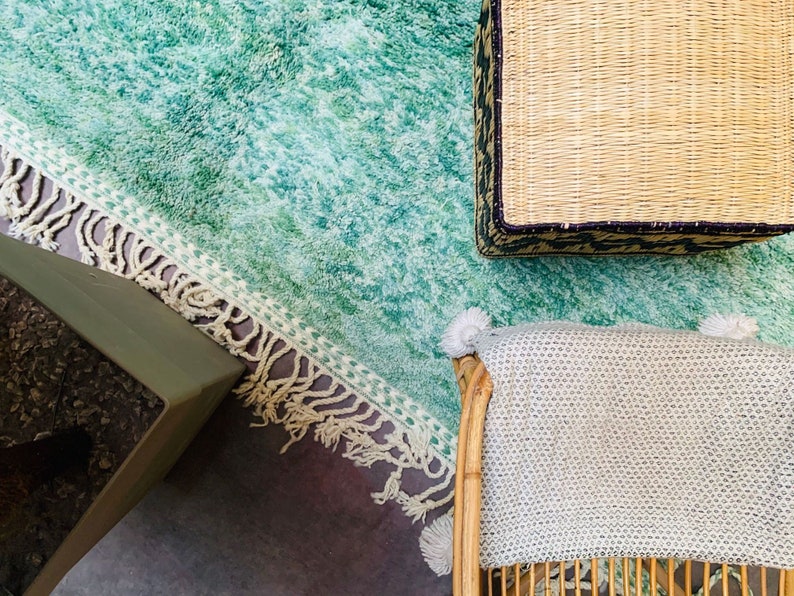 May include: Overhead view of a living room setup featuring a teal shag rug with a fringed border. A woven basket with a dark patterned trim sits atop the rug, along with a light-colored blanket draped over a bamboo chair. A green table is in the corner.