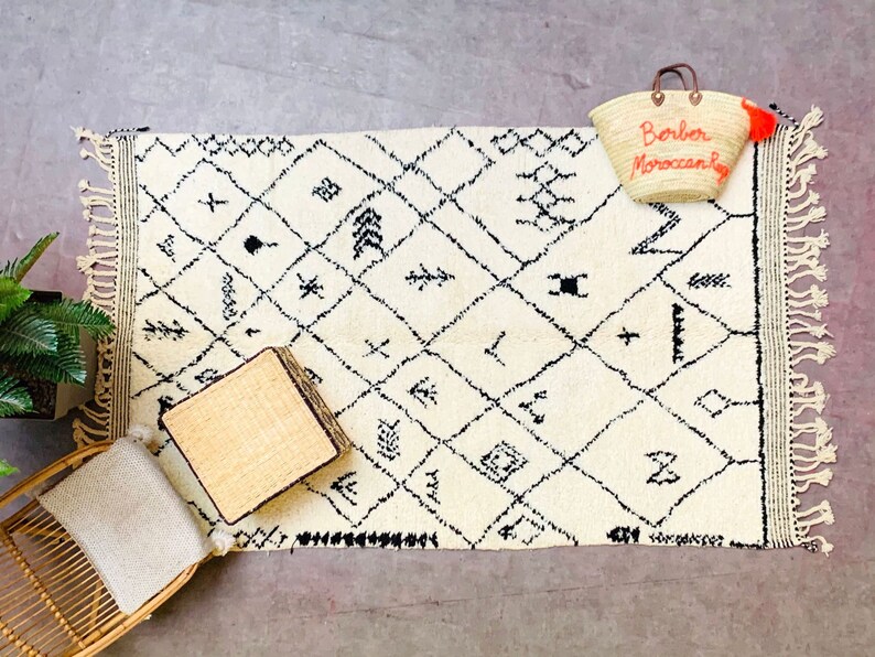 May include: A white and black Moroccan rug with geometric patterns and fringe. A woven basket bag with the text "Borber Moroccan Bag" is on the rug.