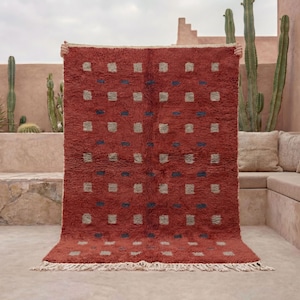 May include: A red rectangular rug with a repeating pattern of square designs in grey and navy blue. The rug has a textured appearance and a fringe along the bottom edge. The background features cacti and a stone wall.