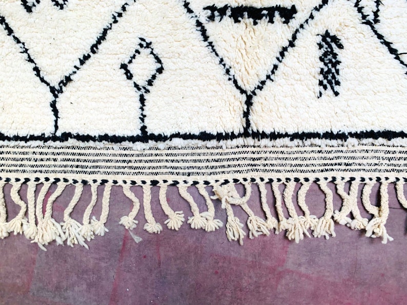 May include: White and black Moroccan rug with a geometric pattern and fringe. The rug has a thick, fluffy pile.