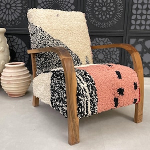 Könnte beinhalten: Ein Holzsessel mit einer einzigartigen, farbenfrohen und gemusterten Stoffpolsterung. Der Stoff zeigt eine Mischung aus weißen, schwarzen und rosafarbenen Farben in einem geometrischen Design. Der Stuhl hat einen dunklen Holzrahmen und Armlehnen.