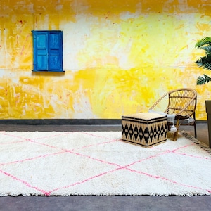 May include: A white shag rug with pink diamond patterns, a woven ottoman, and a wicker chair in front of a yellow wall with a blue window.