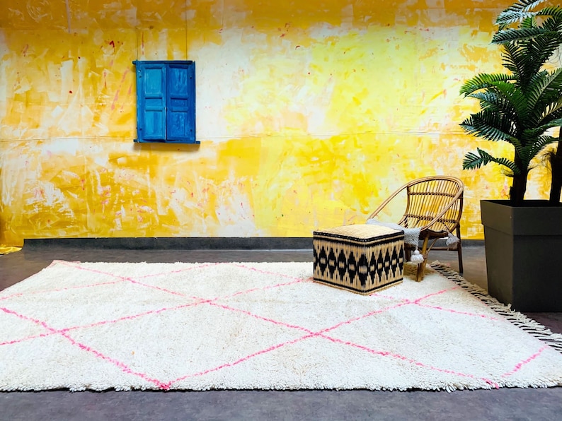 May include: A white shag rug with pink diamond patterns, a woven ottoman, and a wicker chair in front of a yellow wall with a blue window.