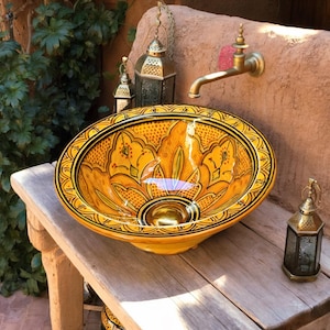 Pode incluir: Uma pia decorativa de cerâmica amarela com um design floral e acabamento preto repousa sobre uma mesa de madeira. Uma torneira de latão e pequenas lanternas são visíveis ao fundo. Um acessório de casa de banho único.