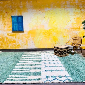 May include: A large, rectangular rug with a teal and white geometric design. The rug features a textured, shaggy pile and a fringe along the edges. The design includes stripes and diamond shapes. The rug is placed on a dark surface against a yellow wall.